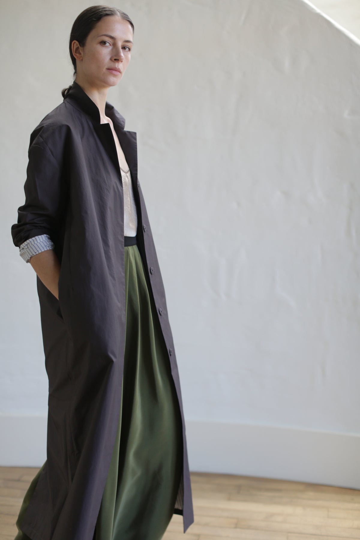 A person stands confidently in a long brown Painter's Coat with one hand in a pocket. They wear a green skirt cinched by a leather Obi Belt and pair it with a light-colored top, posing against a plain cream background.