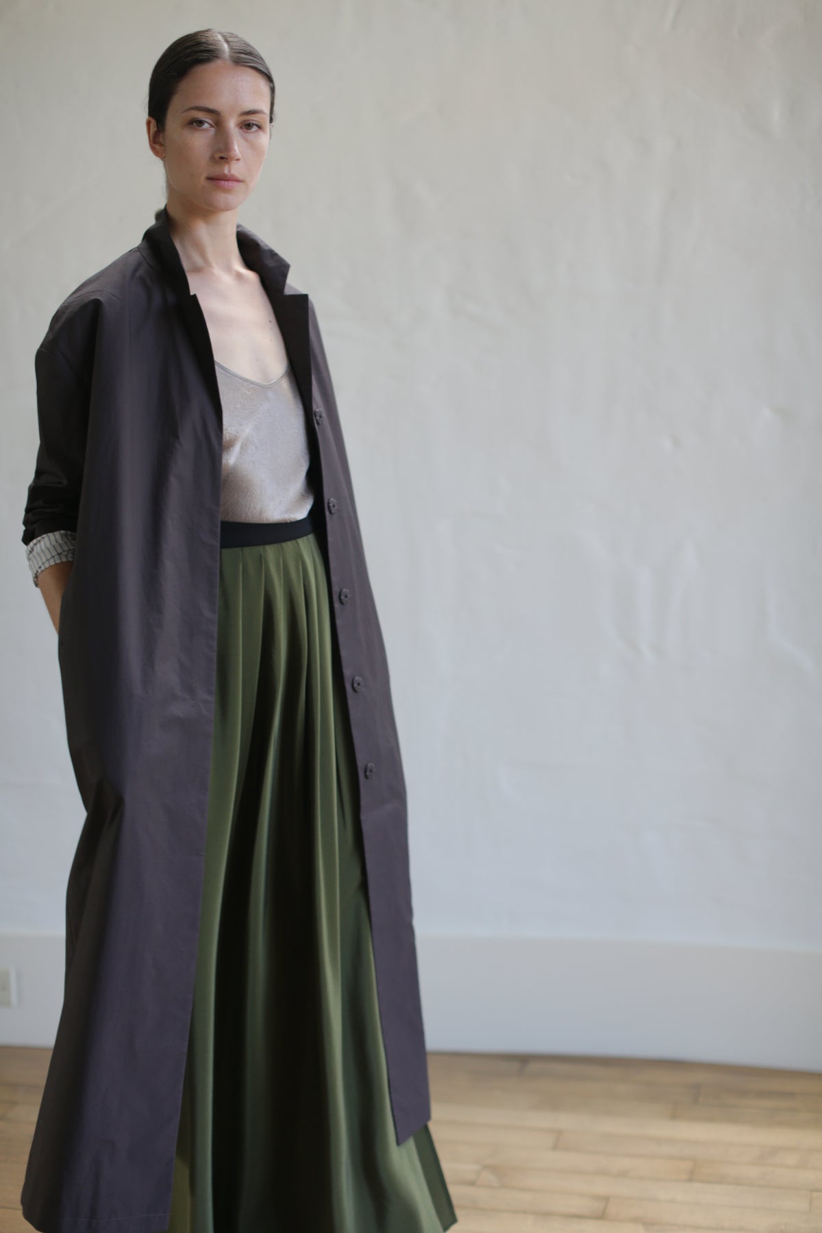 A person stands confidently in a stylish long brown "Painter's Coat," cinched at the waist with a sleek leather Obi belt, paired with a shimmering top and flowing green skirt in a minimalistic room with wooden flooring and plain walls.