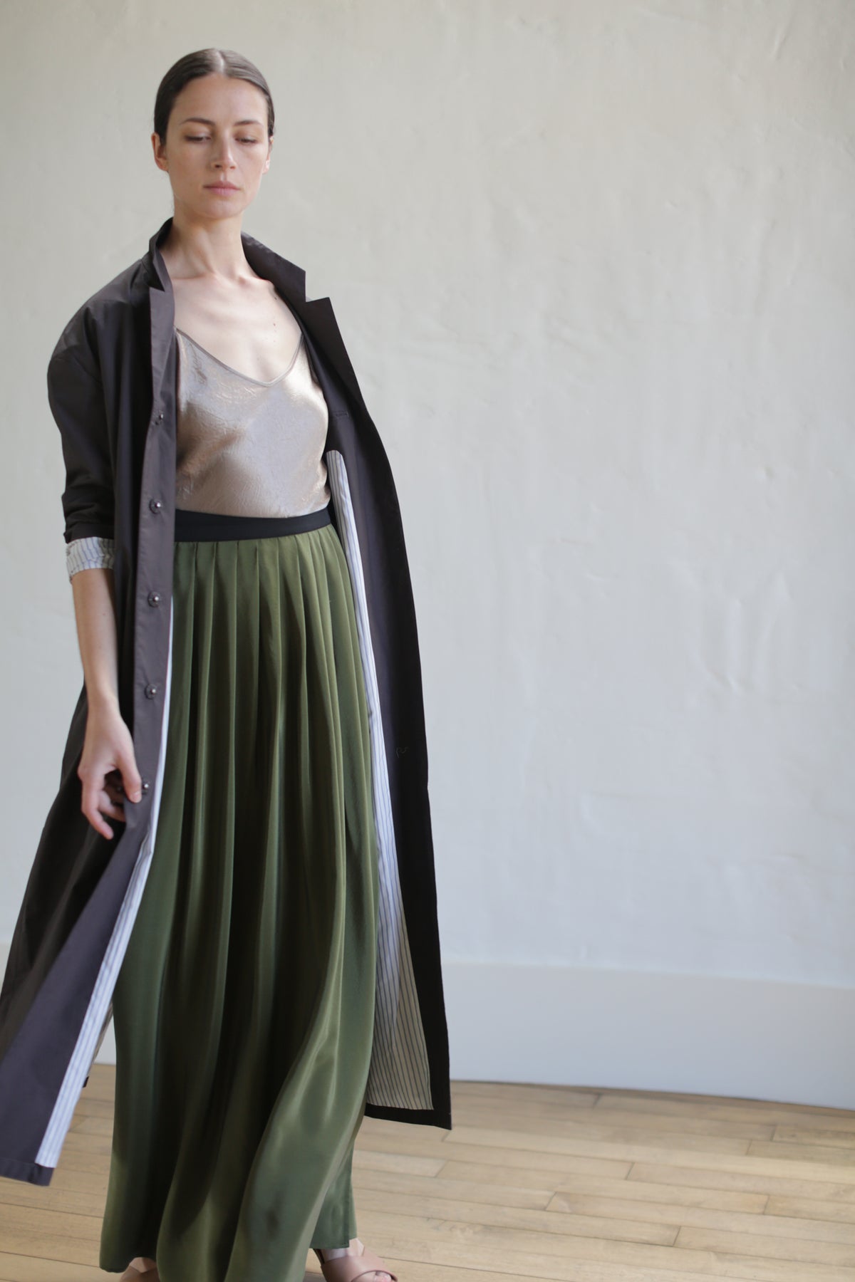 A person stands in a minimalist room, wearing the Painter's Coat in brown over a beige top and a flowing olive green skirt, cinched with a leather obi belt. Their dark hair is pulled back as they look slightly downward, embodying an elegant and contemplative pose.