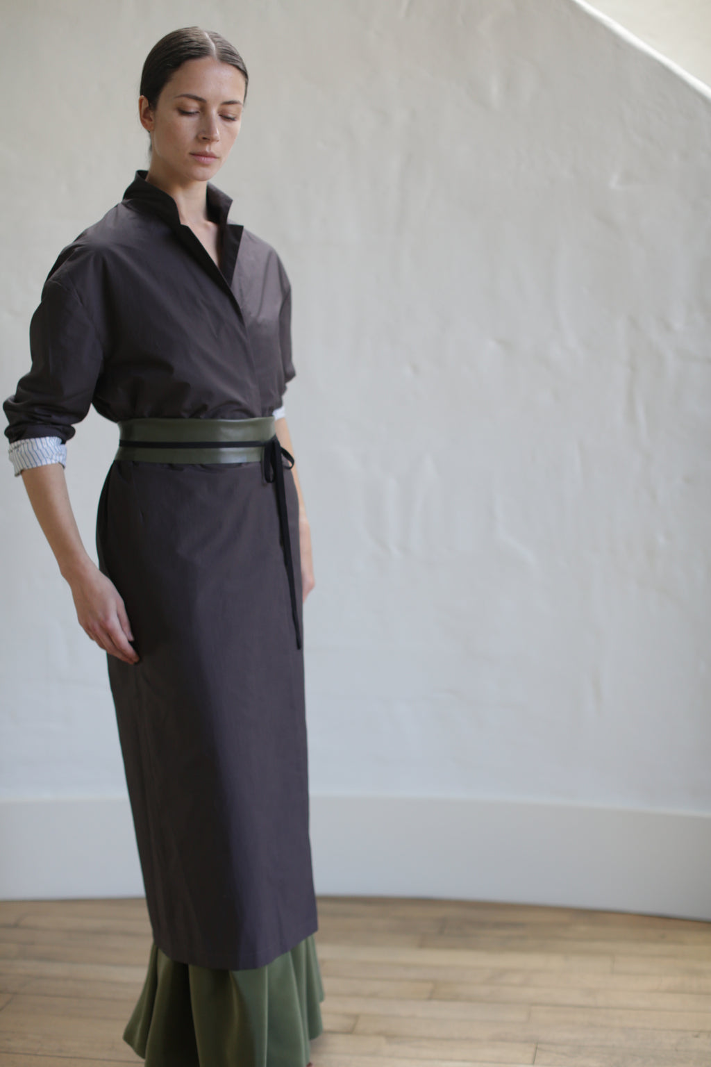 A woman wearing the Brown Painter's Coat with a high collar and a tied leather obi belt stands in a light-filled room with wooden floors and white walls. Her hair is pulled back as she gazes downward, exuding an air of quiet contemplation.