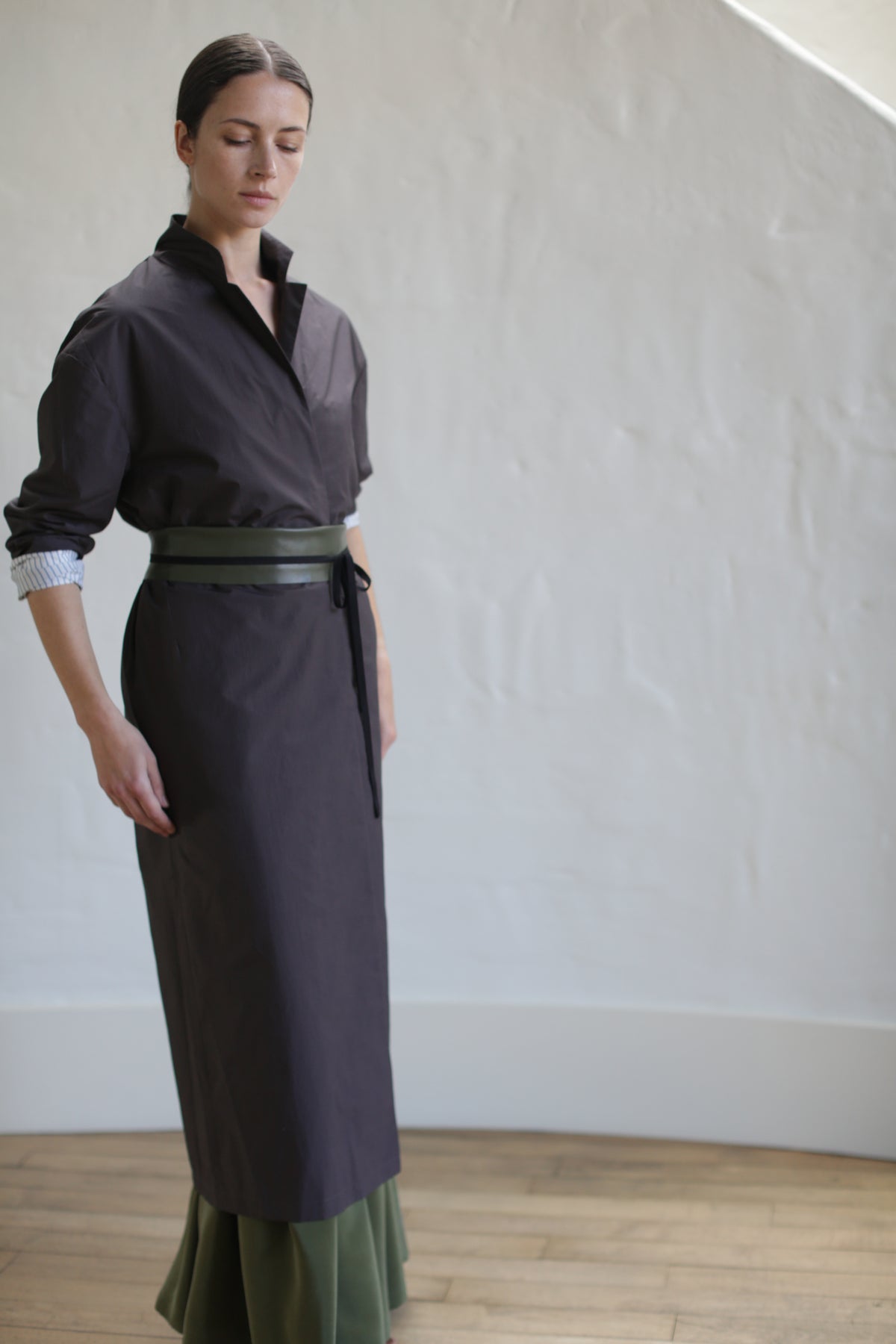 A woman wearing the Brown Painter's Coat with a high collar and a tied leather obi belt stands in a light-filled room with wooden floors and white walls. Her hair is pulled back as she gazes downward, exuding an air of quiet contemplation.