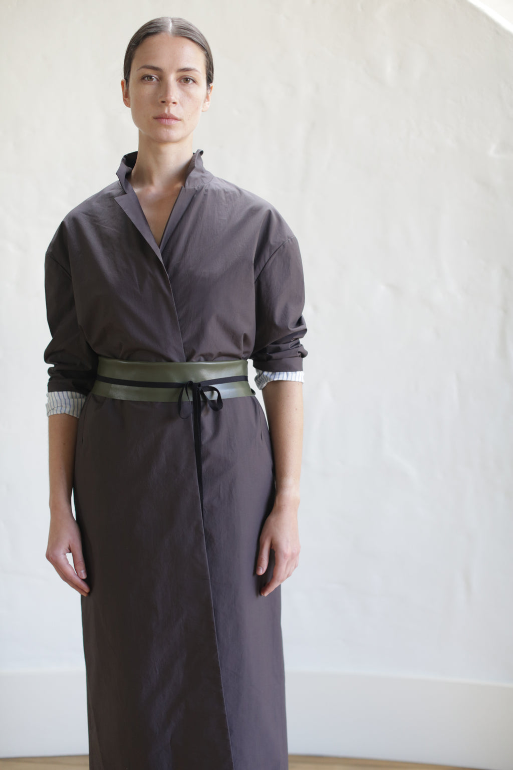 Dressed in a brown Painter's Coat styled like a long robe, cinched with a leather Obi Belt, the individual stands against a light-colored wall. Their hair pulled back, they fix their gaze directly at the camera.