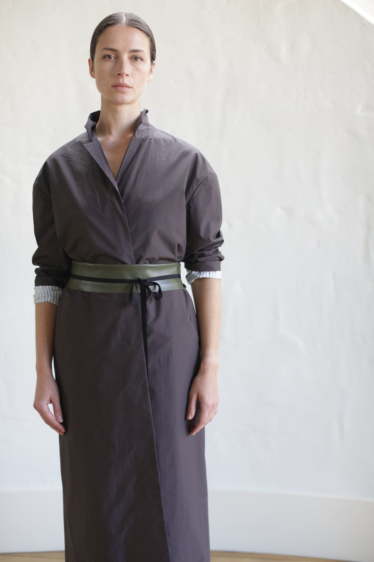 Dressed in a brown Painter's Coat styled like a long robe, cinched with a leather Obi Belt, the individual stands against a light-colored wall. Their hair pulled back, they fix their gaze directly at the camera.