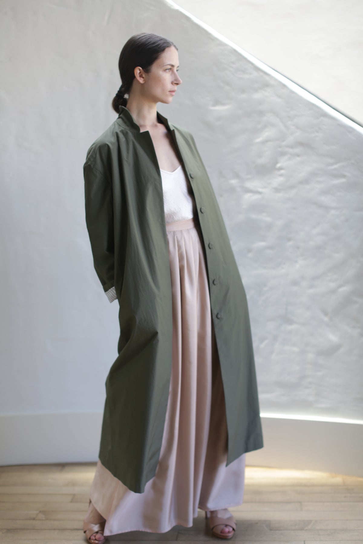 A woman stands indoors before a textured white wall, wearing the Painter's Coat | Olive over a white Typewriter Cotton top and blush pink pants, with her dark hair pulled back.