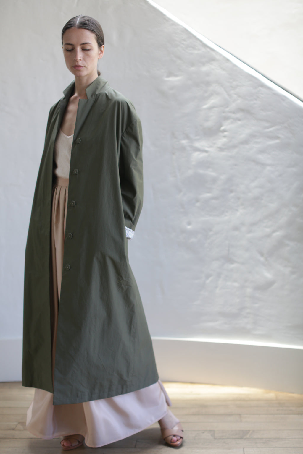 A person stands indoors by a textured white wall, wearing the Painter's Coat in Olive over a beige top and cream wide-legged pants. With brown hair pulled back, they pose in natural light, sandals on their feet.