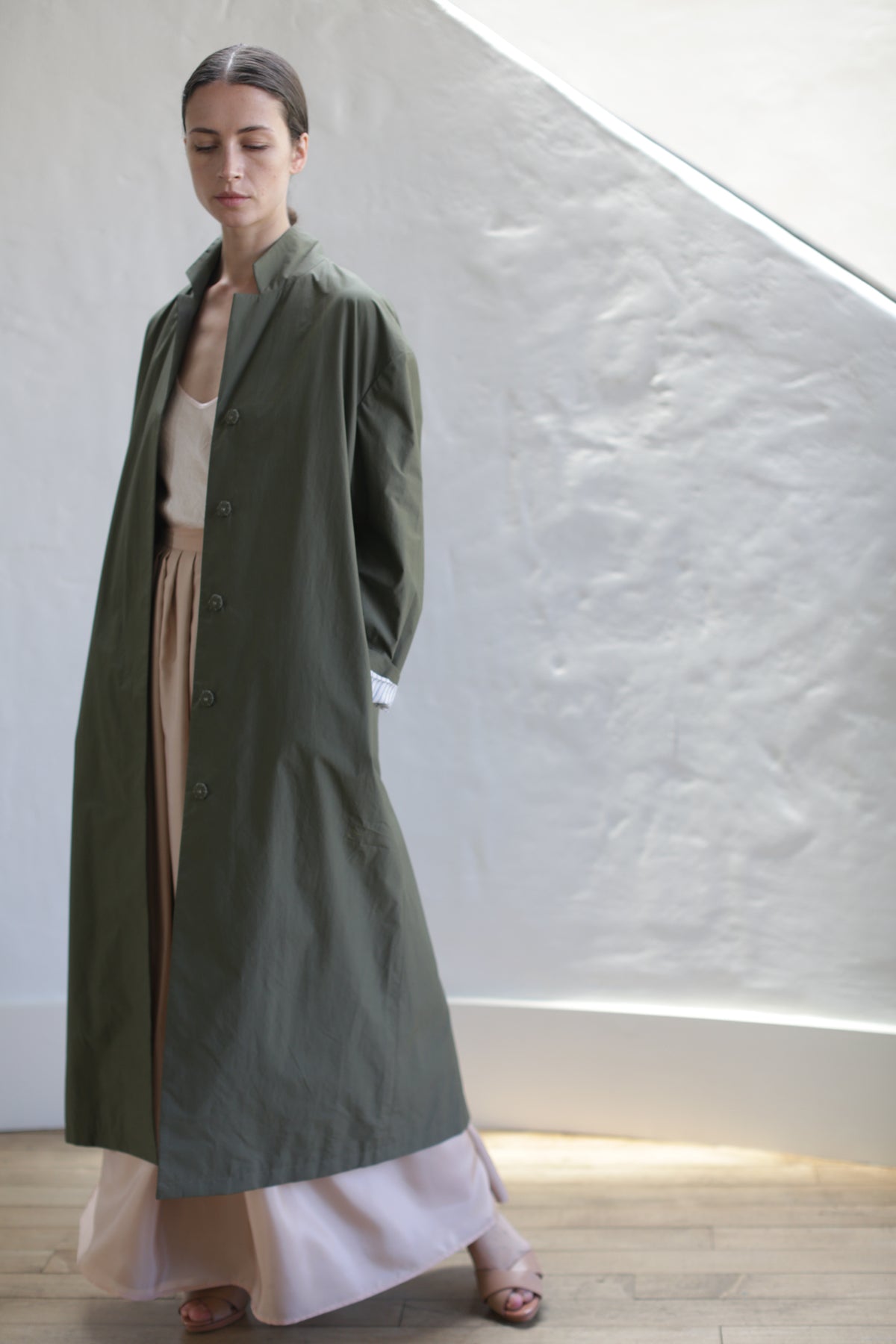 A person stands indoors by a textured white wall, wearing the Painter's Coat in Olive over a beige top and cream wide-legged pants. With brown hair pulled back, they pose in natural light, sandals on their feet.