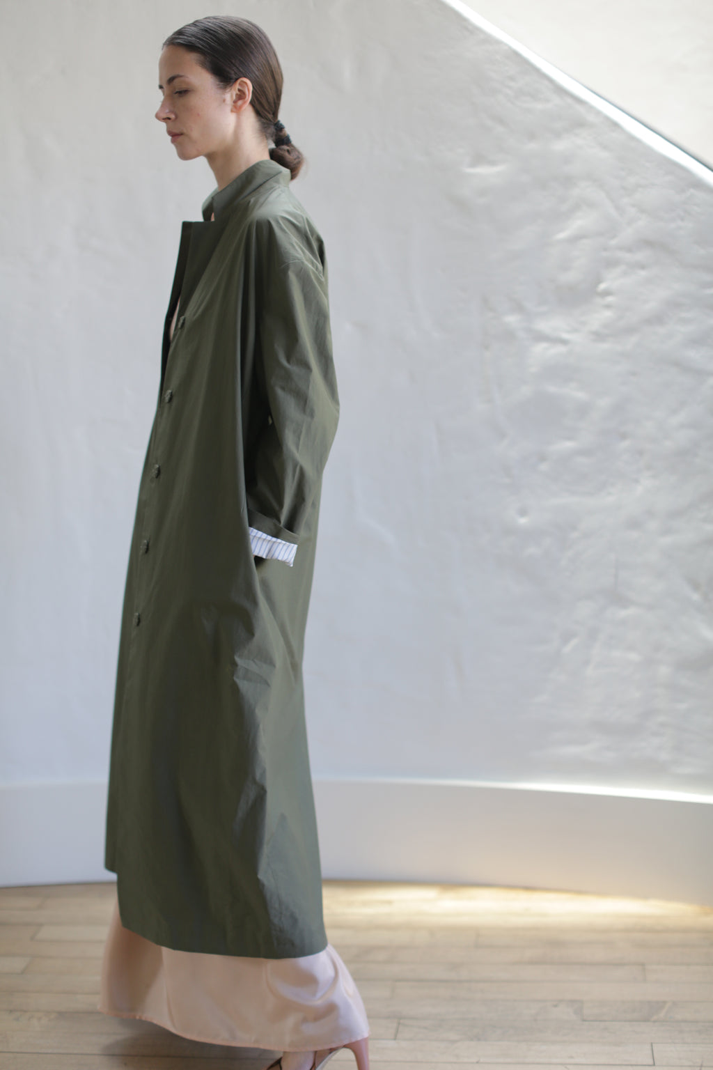 A woman with dark hair tied back wears the Painter's Coat | Olive over a light dress, standing in profile indoors against a textured white wall and wooden floor.