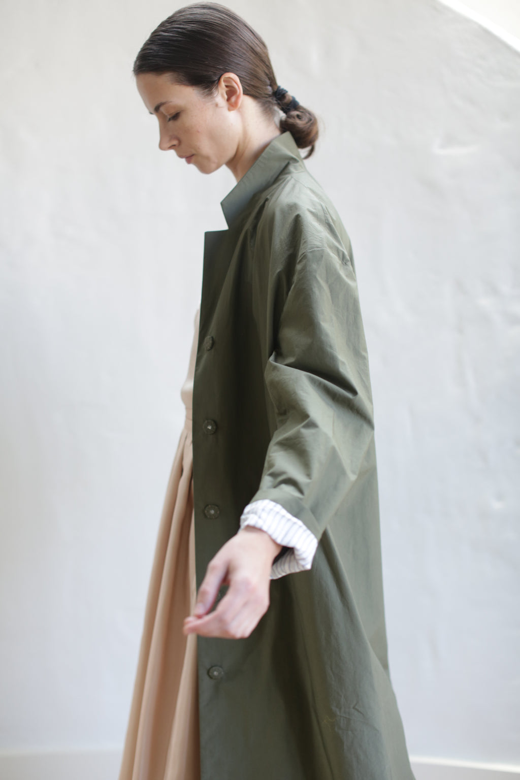 A woman with dark hair tied back stands in profile, wearing the Painter’s Coat | Olive over a light dress, set against a plain light background.