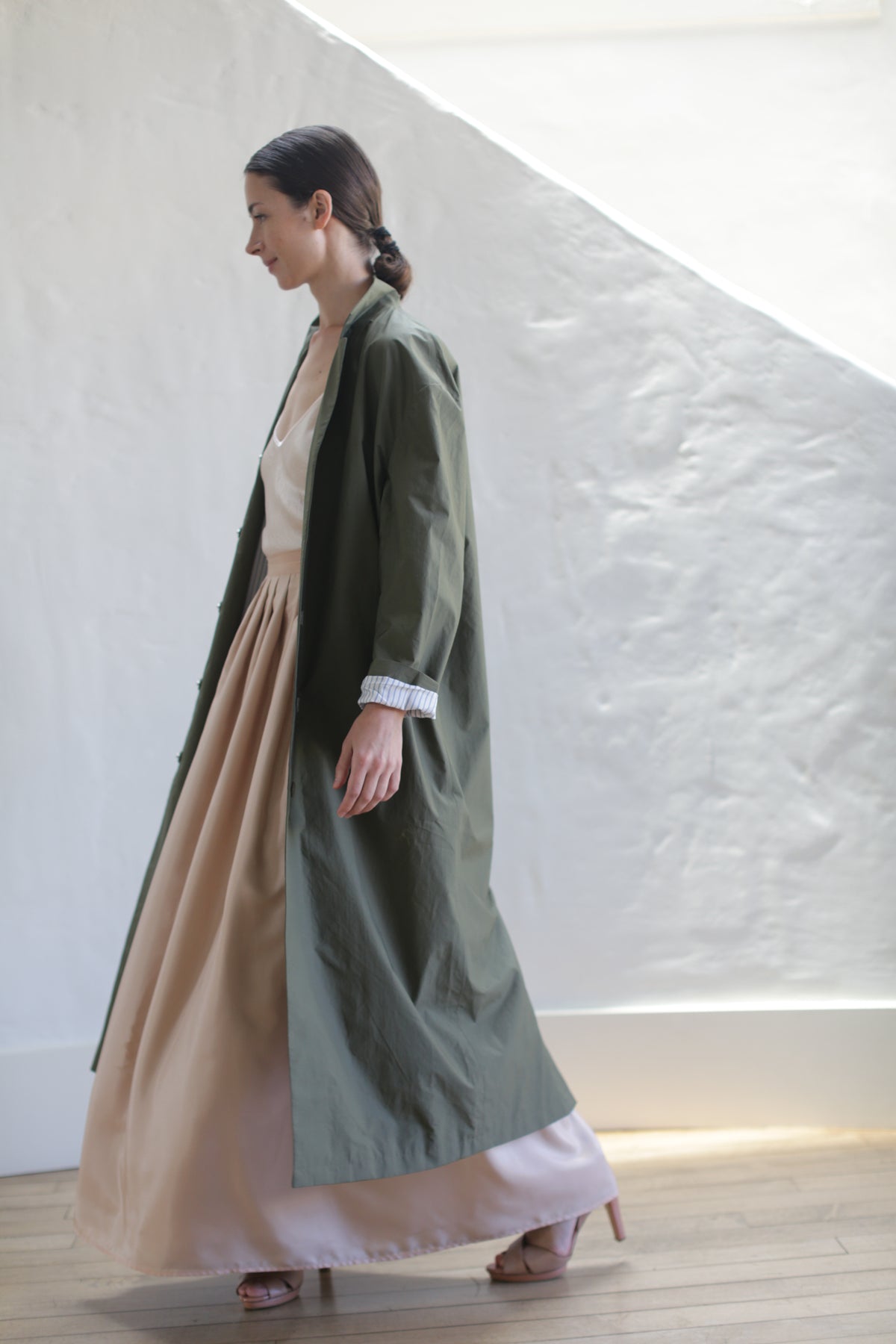 A woman with her hair tied back walks indoors beside a white textured wall, wearing the Painter's Coat | Olive over a flowing beige dress and light pink heels on wooden flooring.