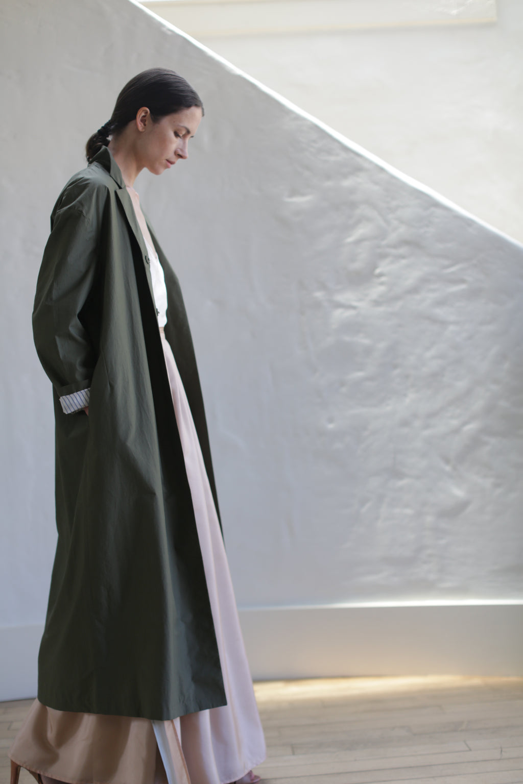 A woman with dark hair tied back, wearing the Painter's Coat | Olive over a light pink dress, stands indoors by a textured white wall as diagonal sunlight casts gentle shadows.