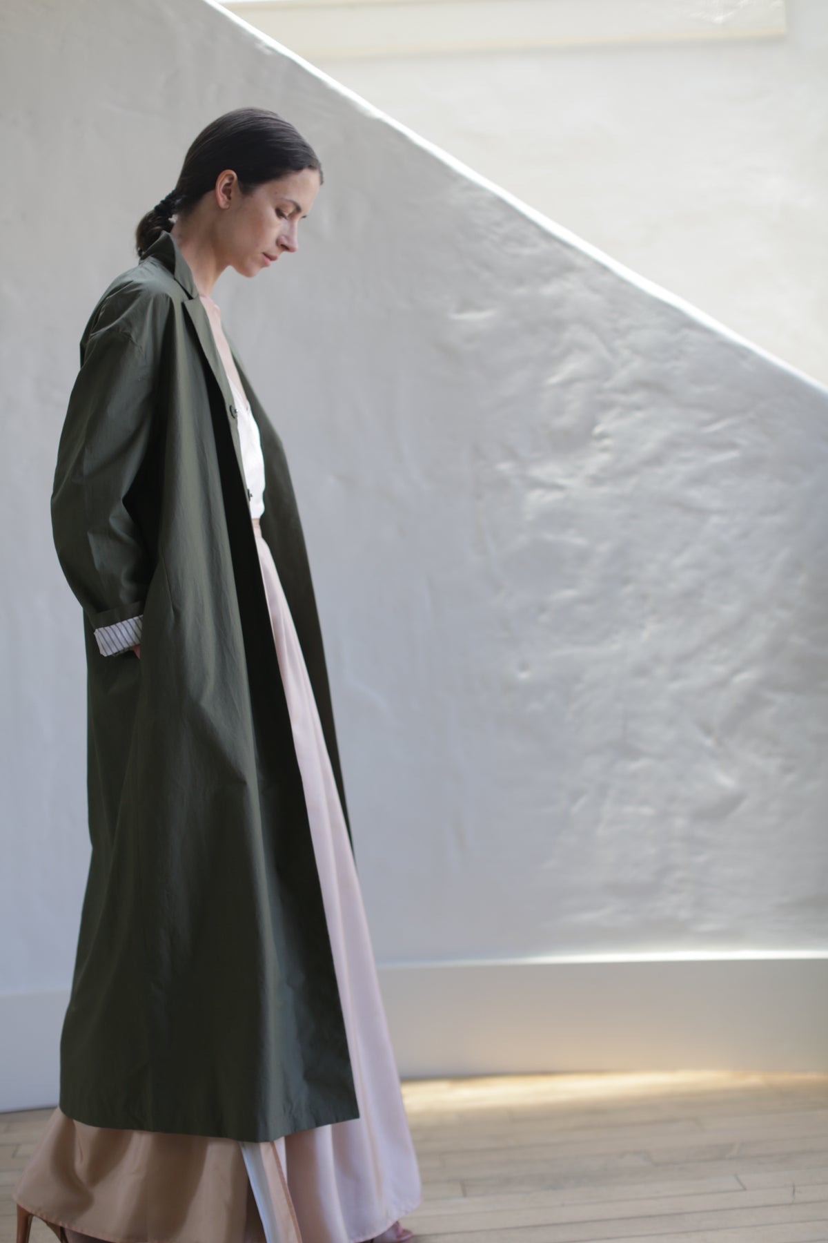 A woman with dark hair tied back, wearing the Painter's Coat | Olive over a light pink dress, stands indoors by a textured white wall as diagonal sunlight casts gentle shadows.
