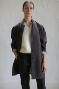 A person with brown hair pulled back is standing against a plain background. They are wearing the Painter's Jacket in dark brown over a white top and black pants, with one hand in a pocket. Their expression is neutral.
