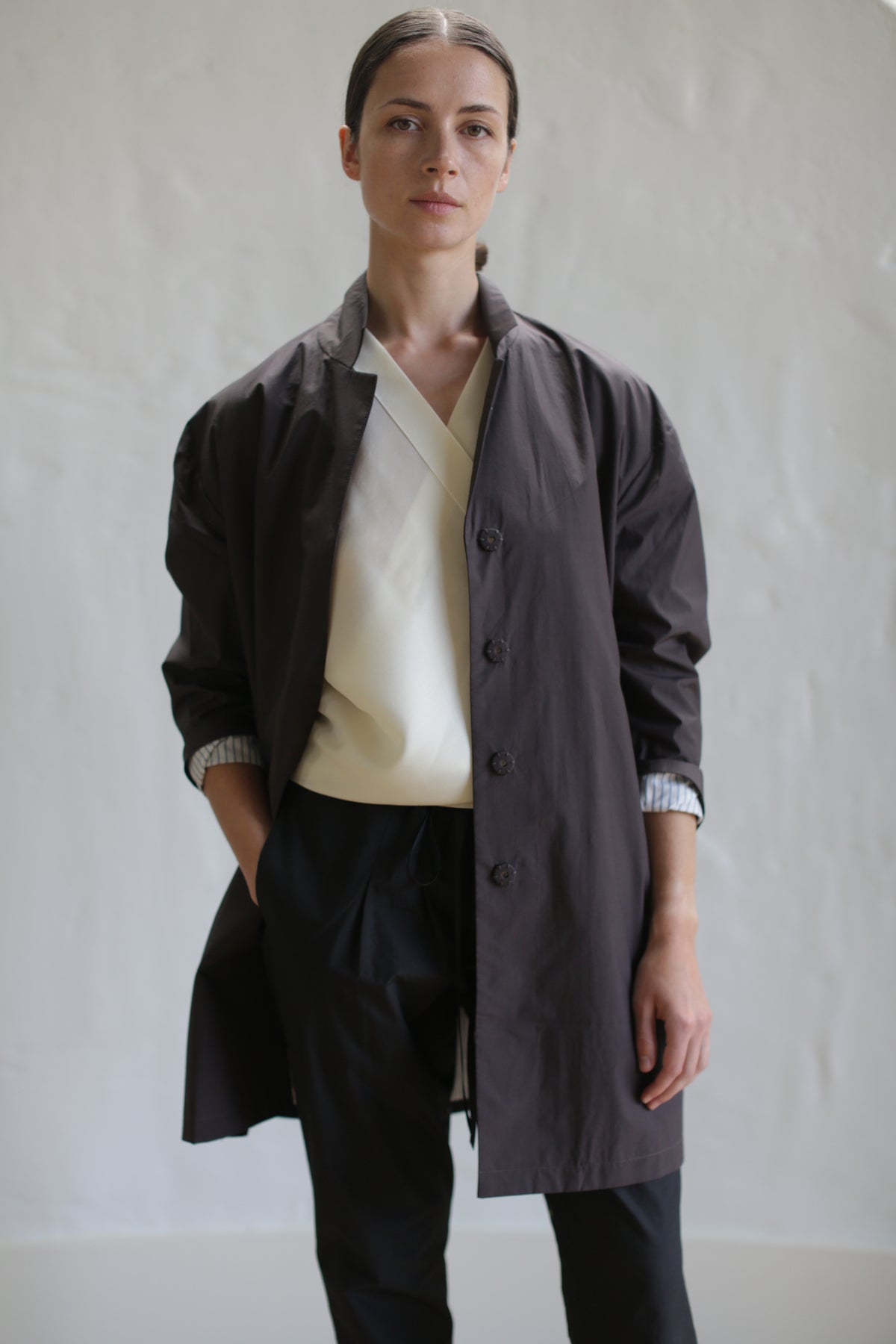 A person with brown hair pulled back is standing against a plain background. They are wearing the Painter's Jacket in dark brown over a white top and black pants, with one hand in a pocket. Their expression is neutral.