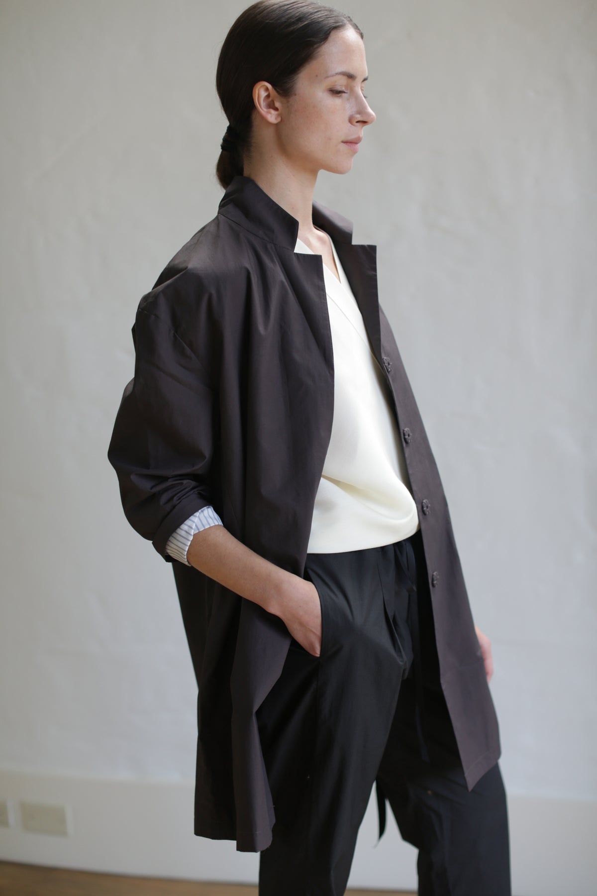 A person with long, dark hair tied back is standing sideways to the camera, wearing a brown Painter's Jacket over a light-colored, tucked-in shirt and dark pants. Their hand is in the jacket pocket as they stand against a plain, light-colored background.