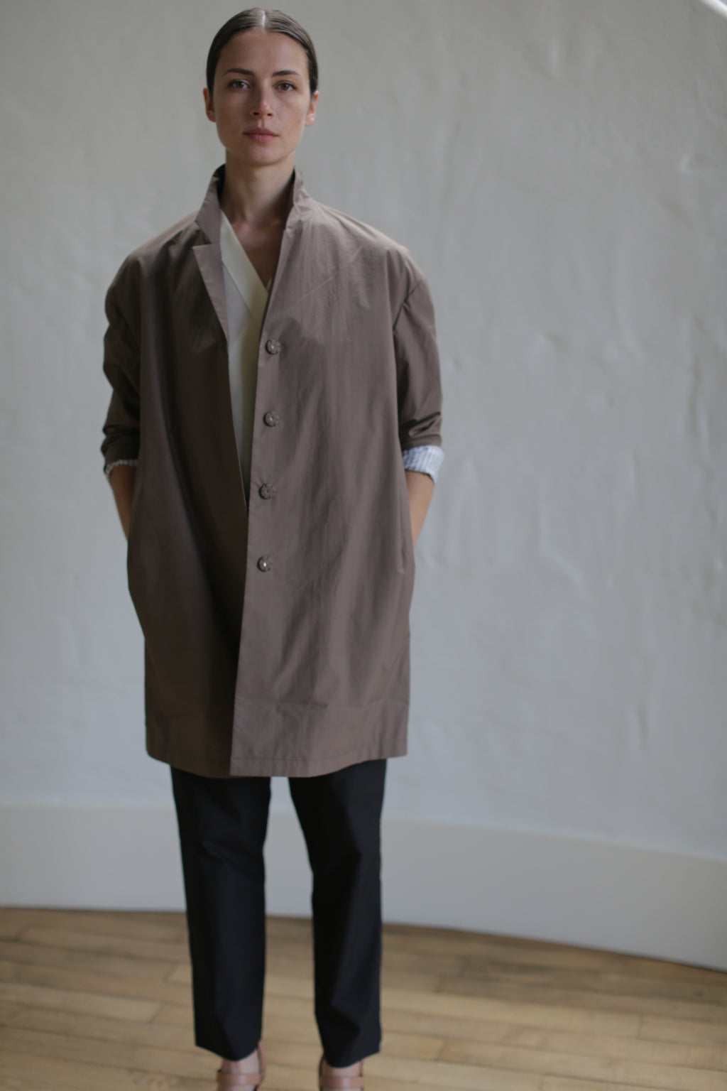 In a room adorned with neutral tones, an individual is wearing a Clay-colored Painter's Jacket over a white shirt and black pants. With hands tucked into the jacket pockets, they gaze directly at the camera, exuding an effortlessly chic style.