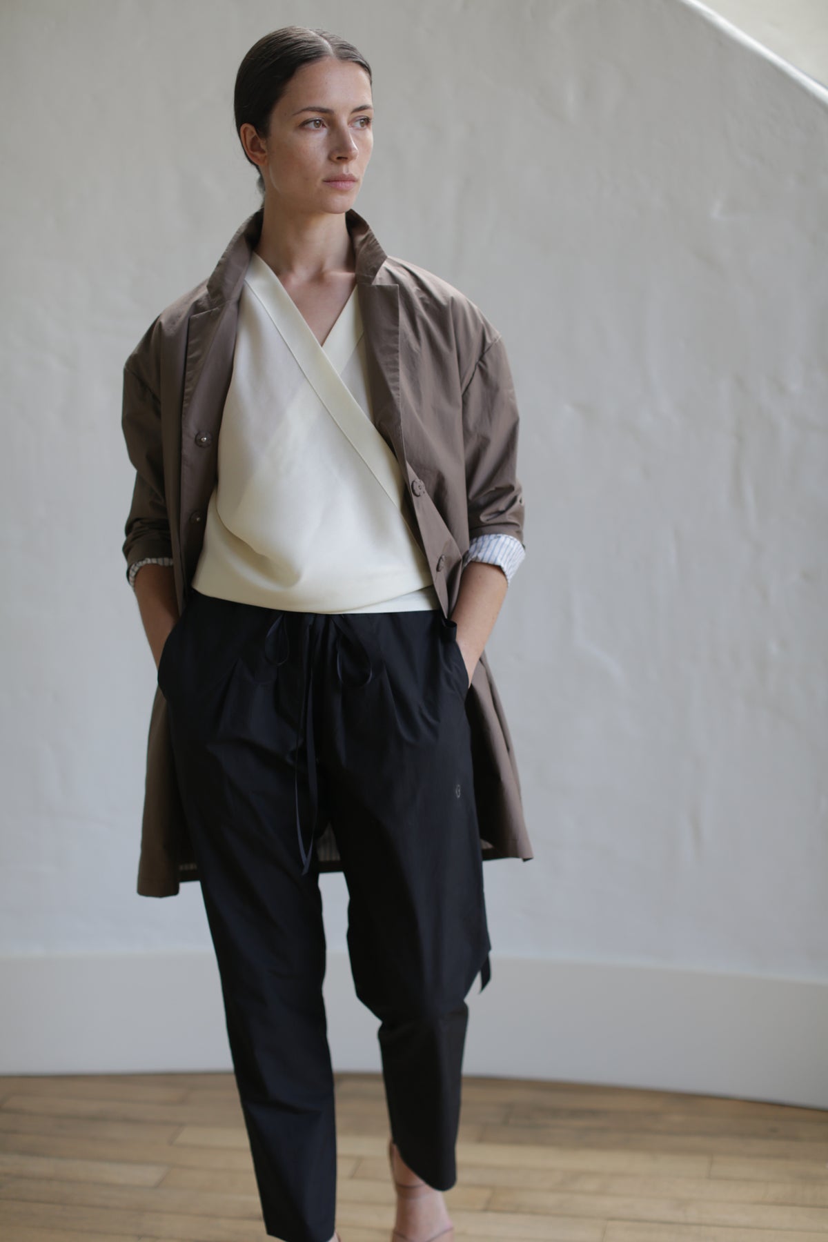 A person stands indoors wearing the Painter's Jacket in Clay over a cream top and black pants. With hands in pockets, they pose against a textured white wall and wooden flooring, exuding an effortless style.