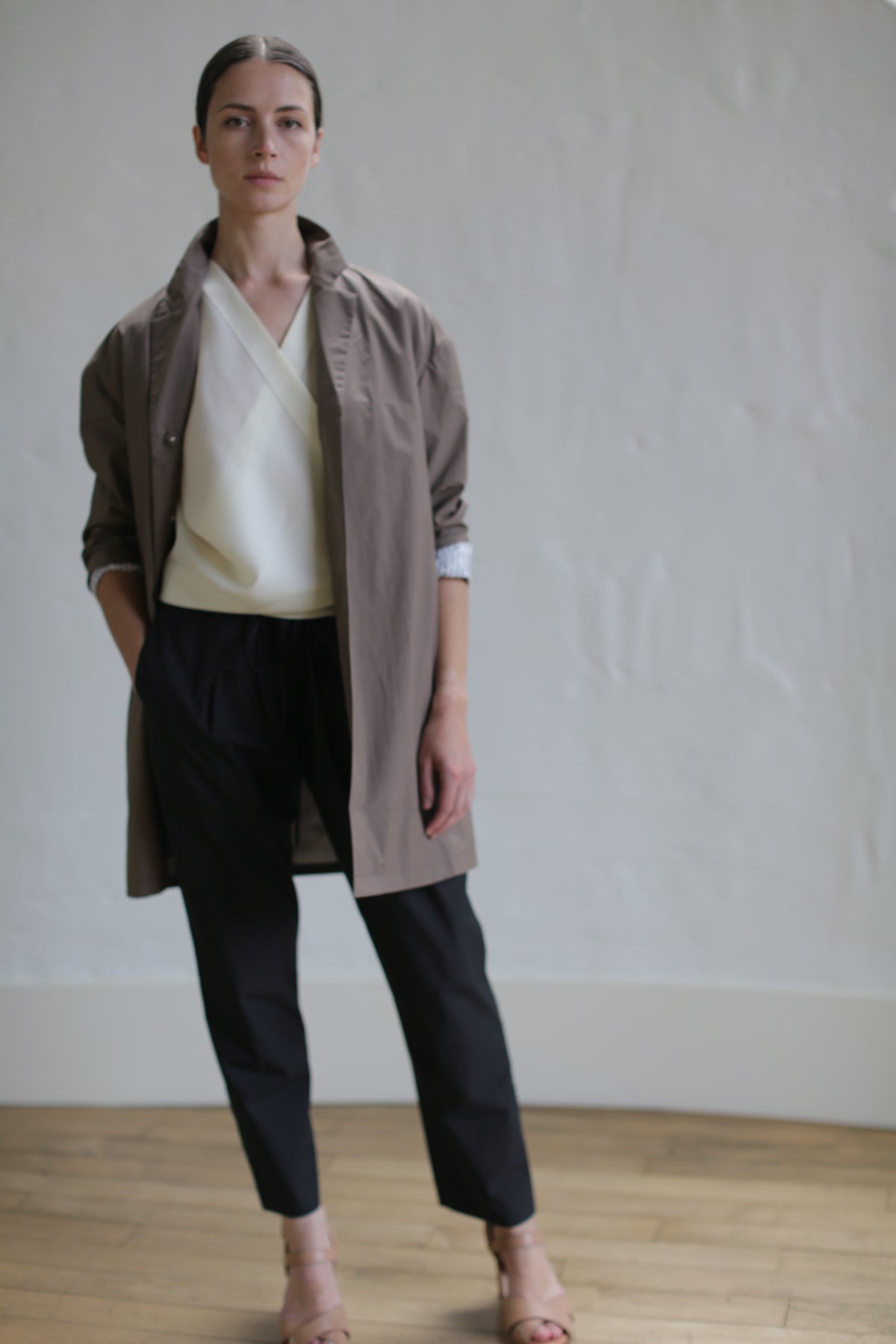 A person stands indoors against a plain wall, wearing a light-colored blouse, a Painter's Jacket in Clay, and black pants. A hand rests in their pocket. The wooden floor and soft lighting add to the minimalistic setting.