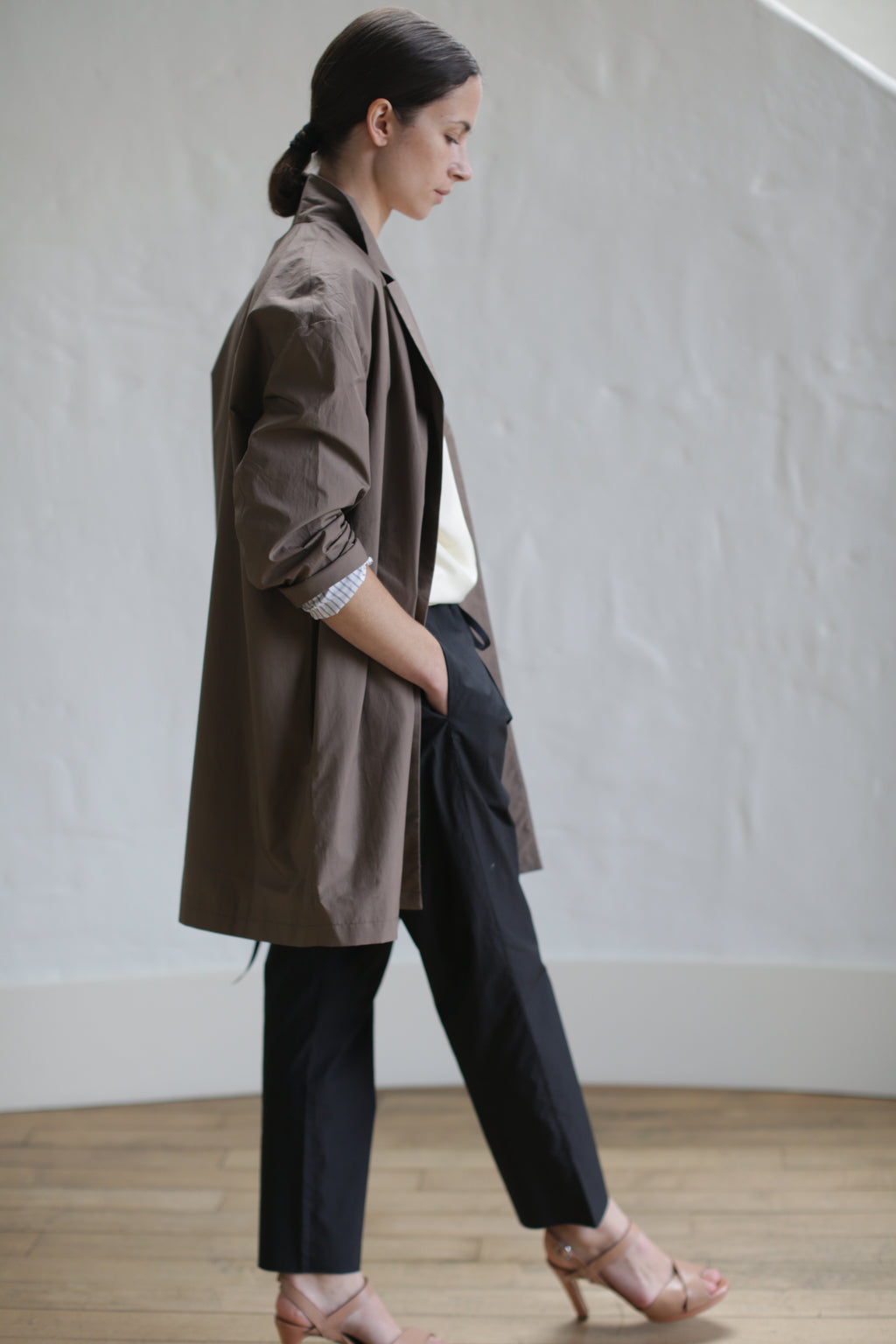 A person stands in profile against a light background, wearing a clay-colored Painter's Jacket, dark pants, and beige high heels. Their hair is tied back, and one hand is in their pocket. The setting appears to be indoors with a wooden floor.