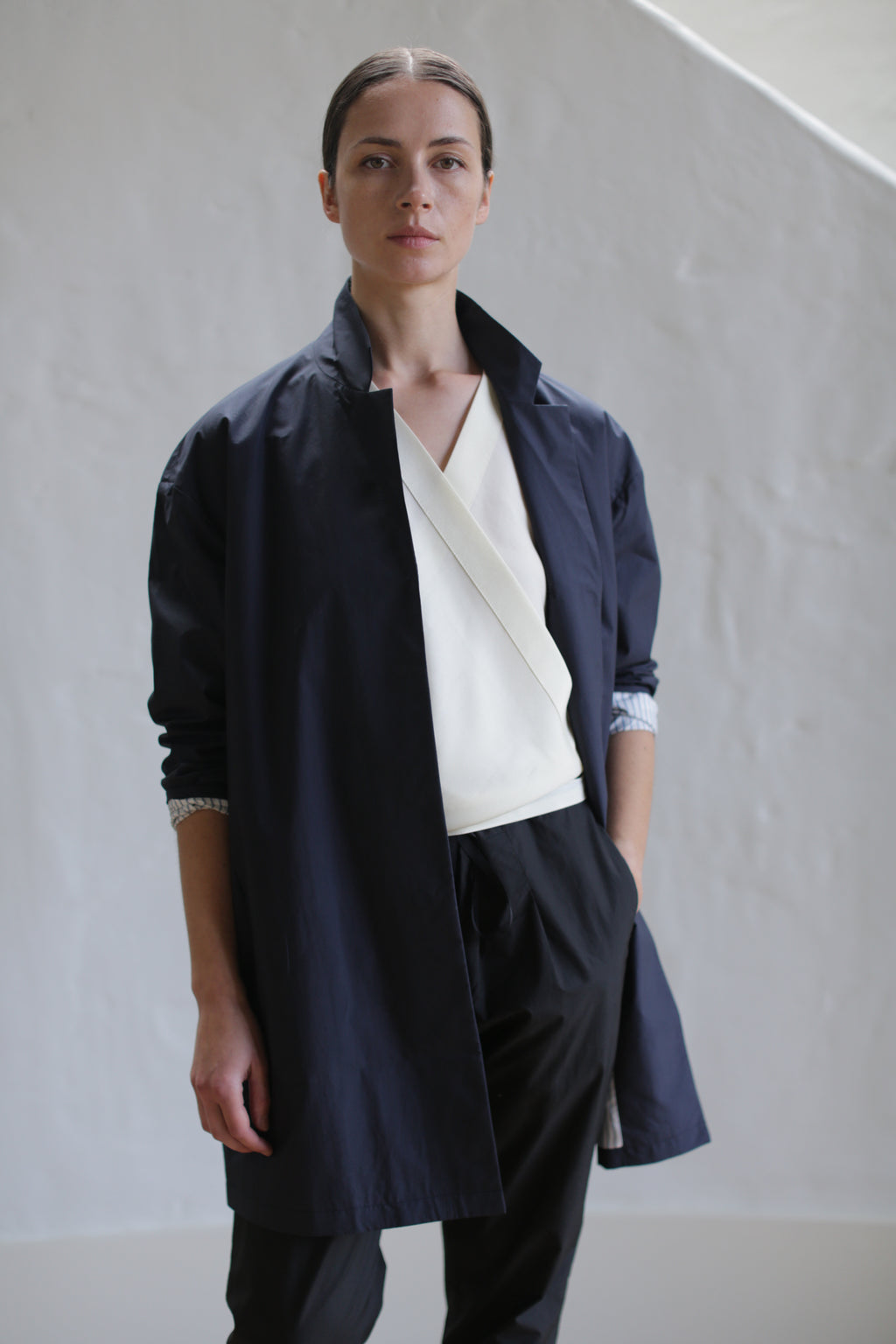 A person stands against a textured white wall, wearing the Painter's Jacket in Navy over a cream-colored top with a layered design. Their dark pants match the jacket, and their hair is neatly pulled back. A calm expression graces their face.