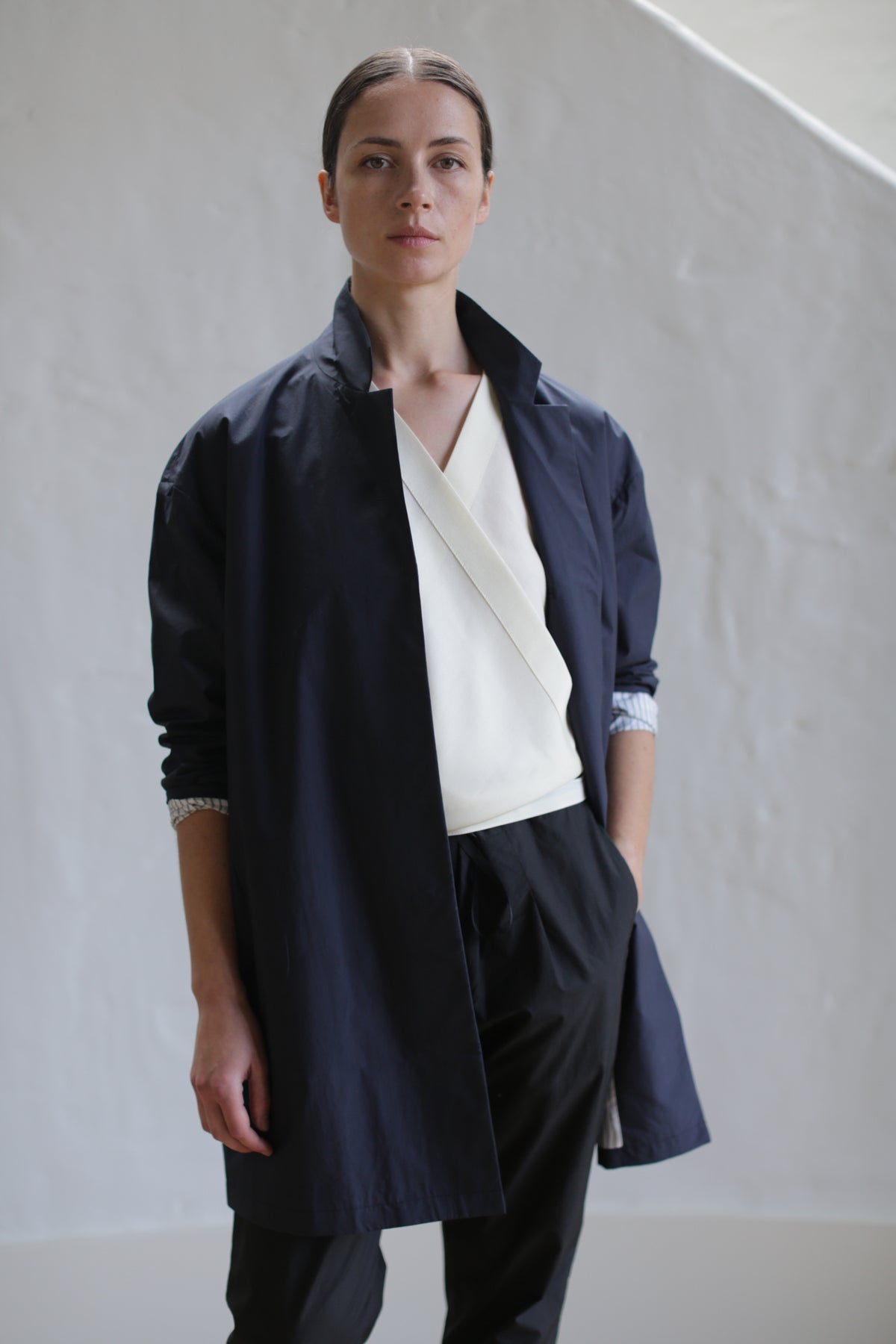 A person stands against a textured white wall, wearing the Painter's Jacket in Navy over a cream-colored top with a layered design. Their dark pants match the jacket, and their hair is neatly pulled back. A calm expression graces their face.