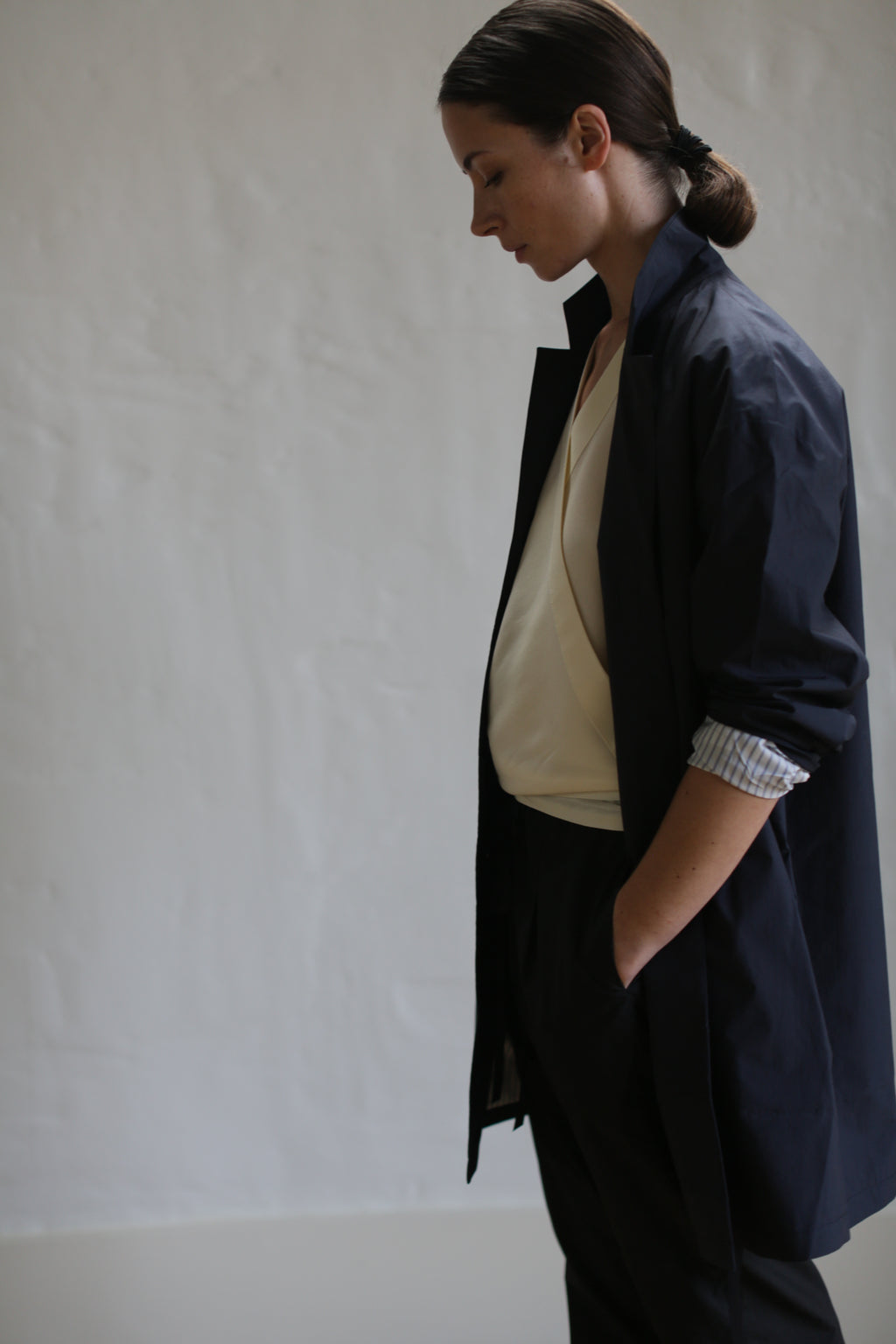 A person with dark hair styled in a low bun is wearing the Painter's Jacket in Navy, which is long and evocative of a painter's coat, over a light top. They stand sideways, looking down, with one hand in their pocket against a plain background.