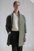 A person with a neutral expression stands with one hand in their pocket, wearing the Painter's Jacket in Olive over a white top and black pants. The background is a plain, light-colored wall.