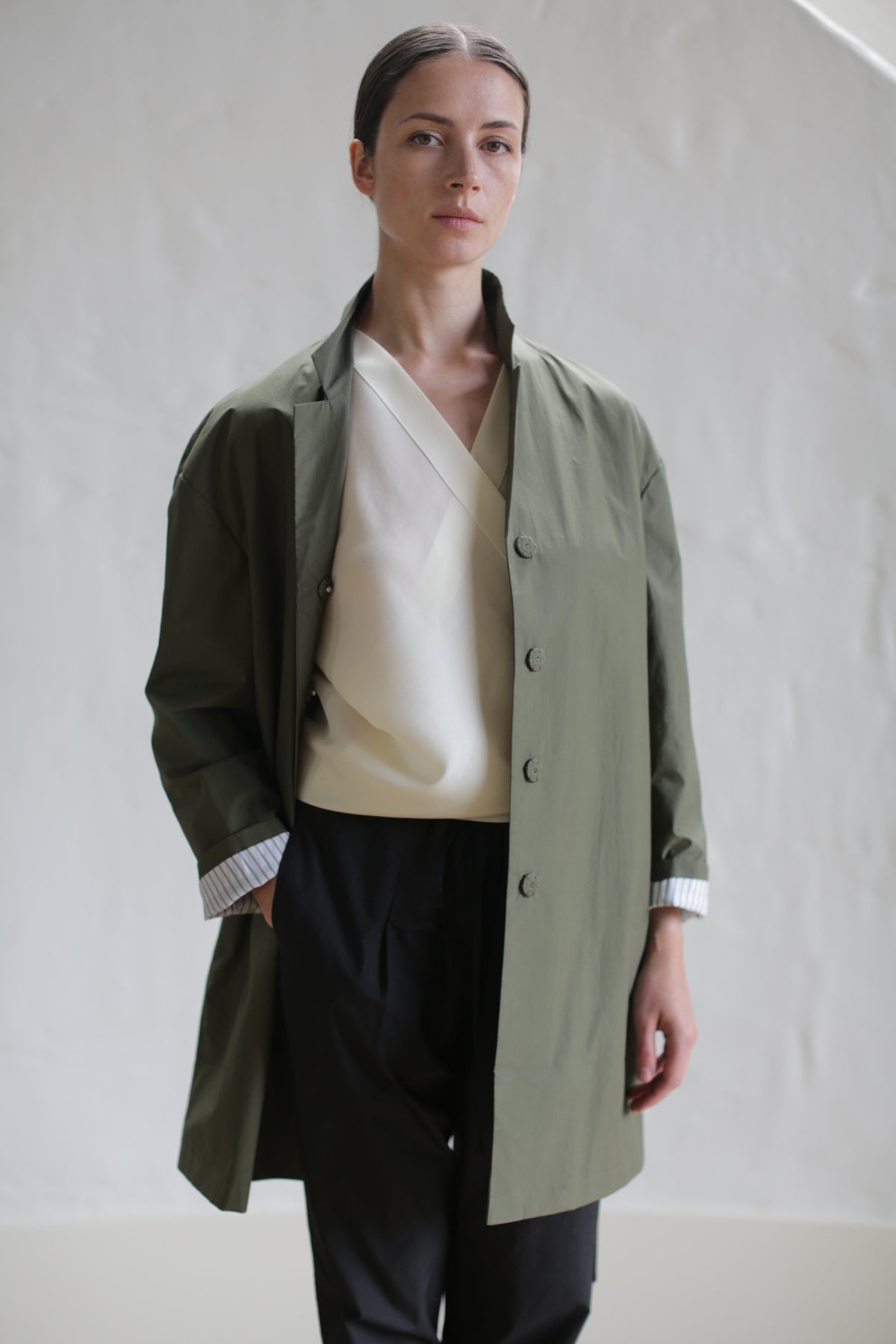 A person with a neutral expression stands with one hand in their pocket, wearing the Painter's Jacket in Olive over a white top and black pants. The background is a plain, light-colored wall.
