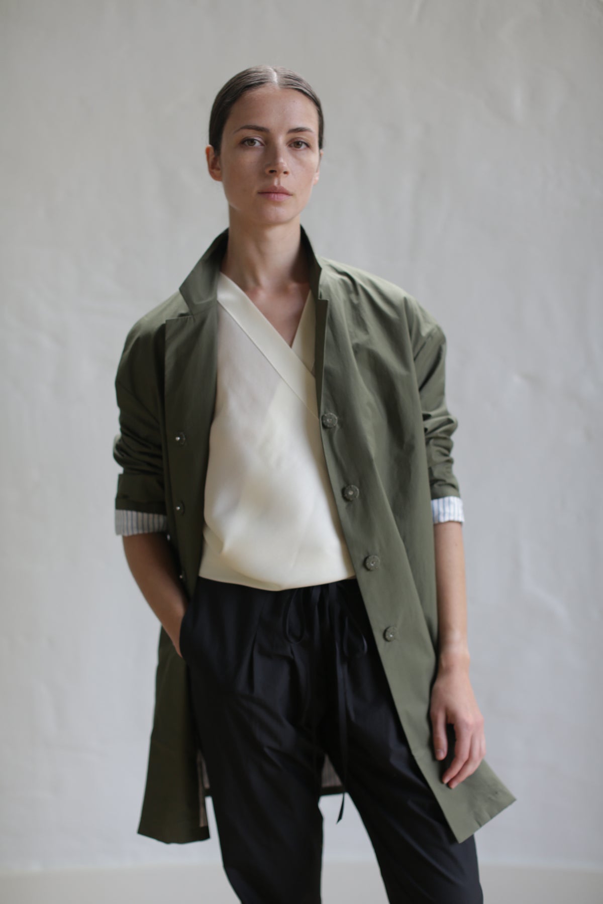 A person stands in front of a plain background, wearing the Painter's Jacket in Olive over a white V-neck top and black trousers. The person's hair is tied back in a sleek, low bun, and their expression is neutral.
