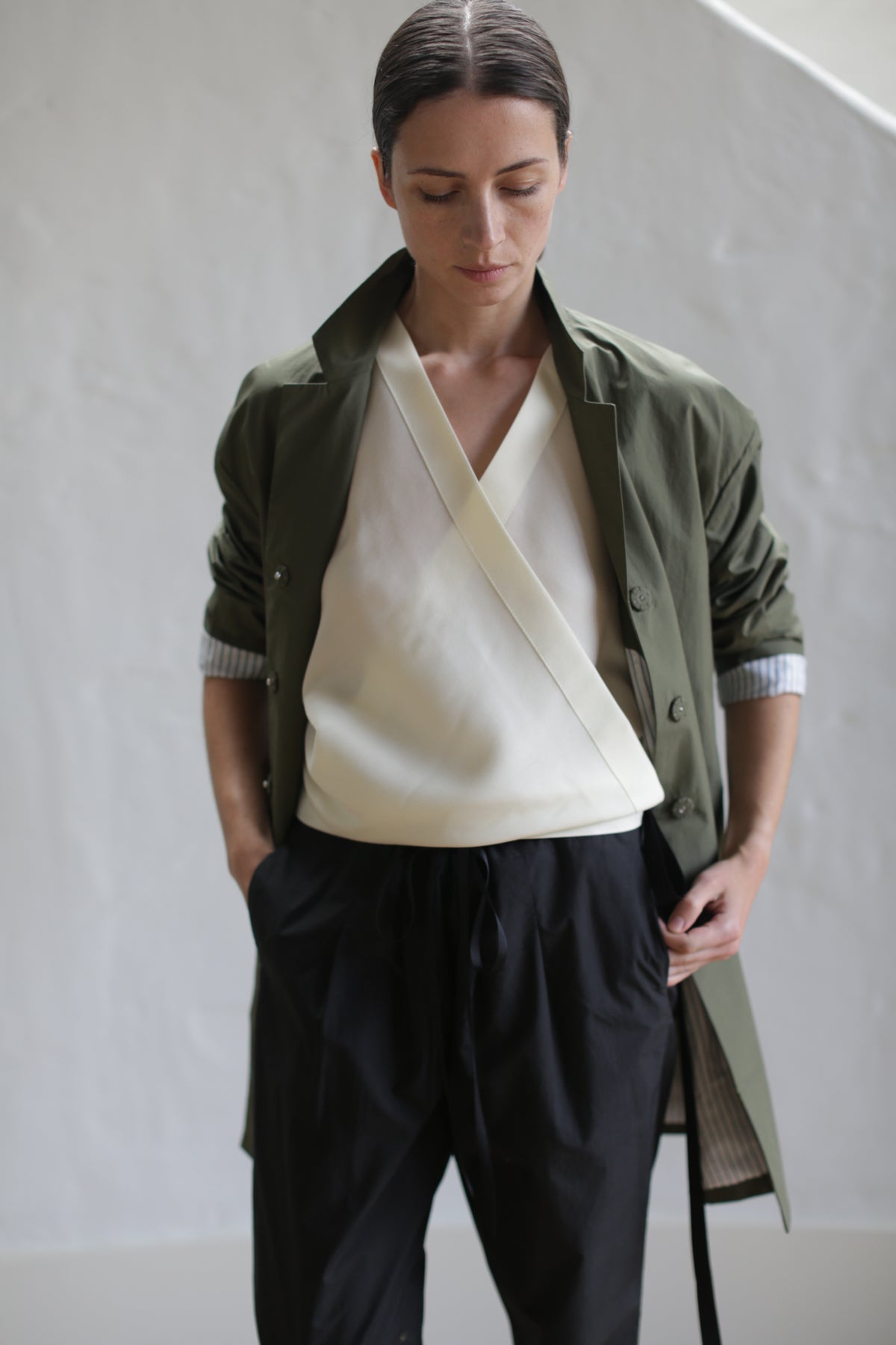 A person with dark hair tied back stands with hands in pockets, wearing an olive Painter's Jacket, a white wrap blouse, and black pants with a leather obi belt. The individual looks downward in a minimalist, light-colored indoor setting.