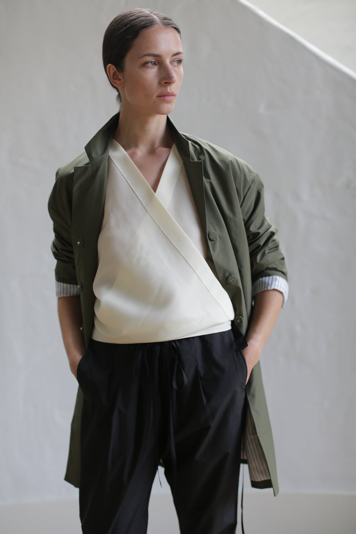 A person with neatly tied back hair stands confidently, wearing a light beige wrap top cinched with a leather Obi Belt, loose black trousers, and the Painter's Jacket in olive with rolled-up sleeves. The individual has their hands in their pockets and is posing against a neutral-colored background.