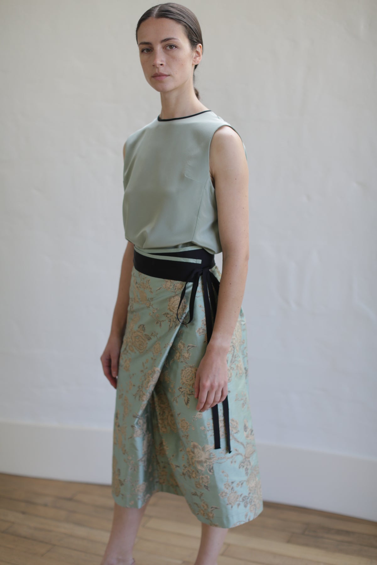 A person wearing a sleeveless, light green top tucked into the limited edition Dupioni Petal Wrap Skirt in Jade stands against a plain wall. The outfit showcases pastel colors and an elegant, zero-waste design. The wooden floor contrasts with the neutral background.