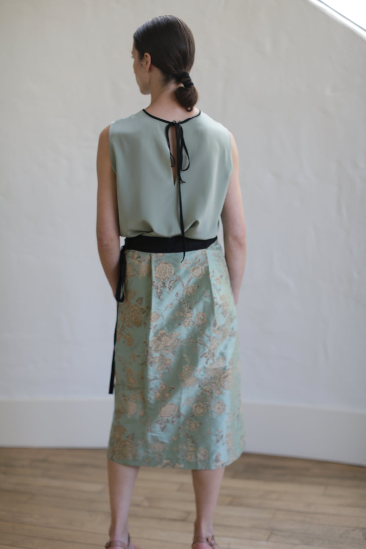 A person in a sleeveless light green top made of Dupioni silk with a bow at the back, paired with the limited edition Dupioni Petal Wrap Skirt in Jade. They face away on a wooden floor beside a textured white wall, highlighting their exquisite style.
