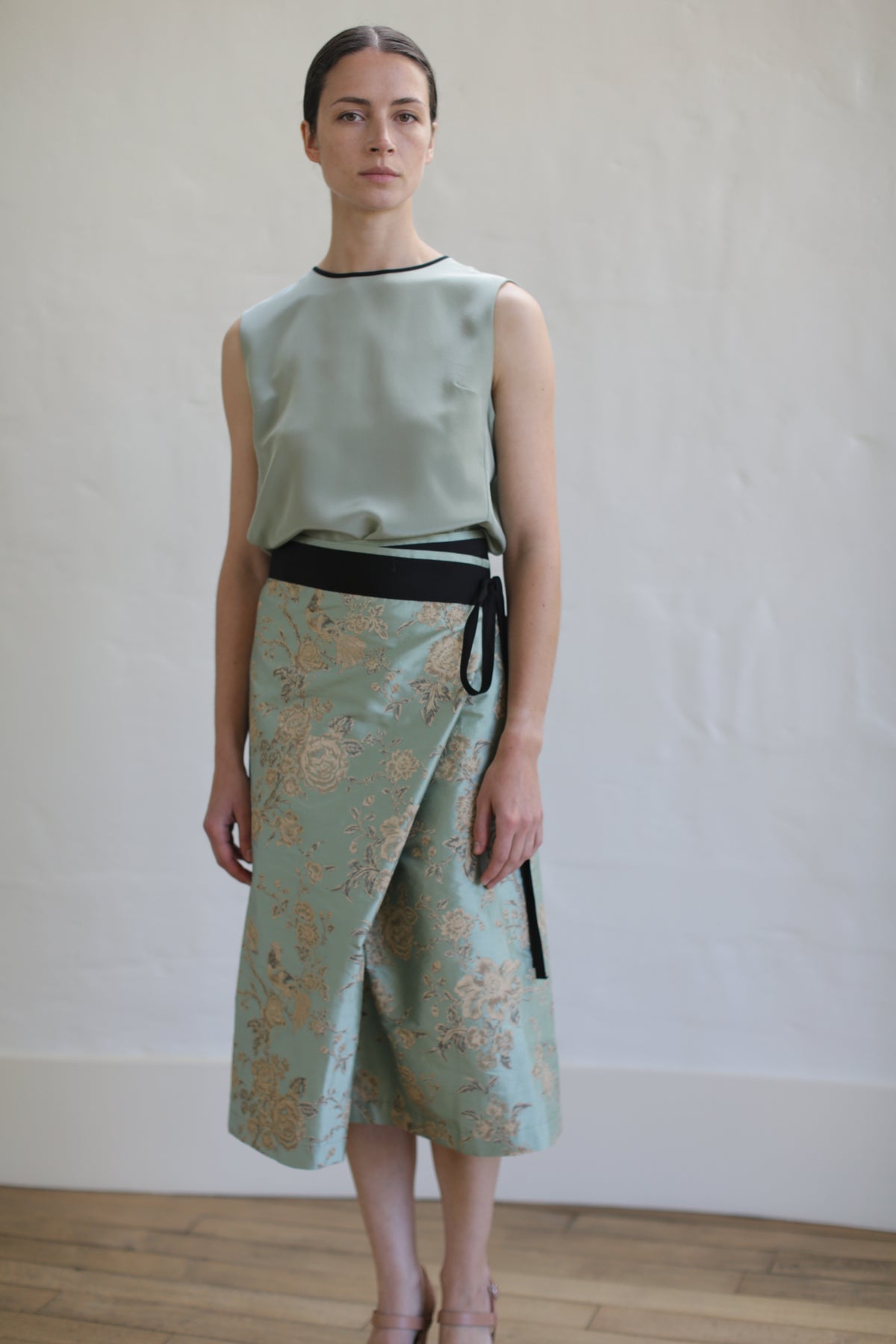 A person stands against a light-colored wall, wearing a sleeveless light green top paired with the Dupioni Petal Wrap Skirt in Jade, adorned with floral patterns. A black ribbon accents the waist. The wooden floor enhances the setting's classic and elegant vibe.
