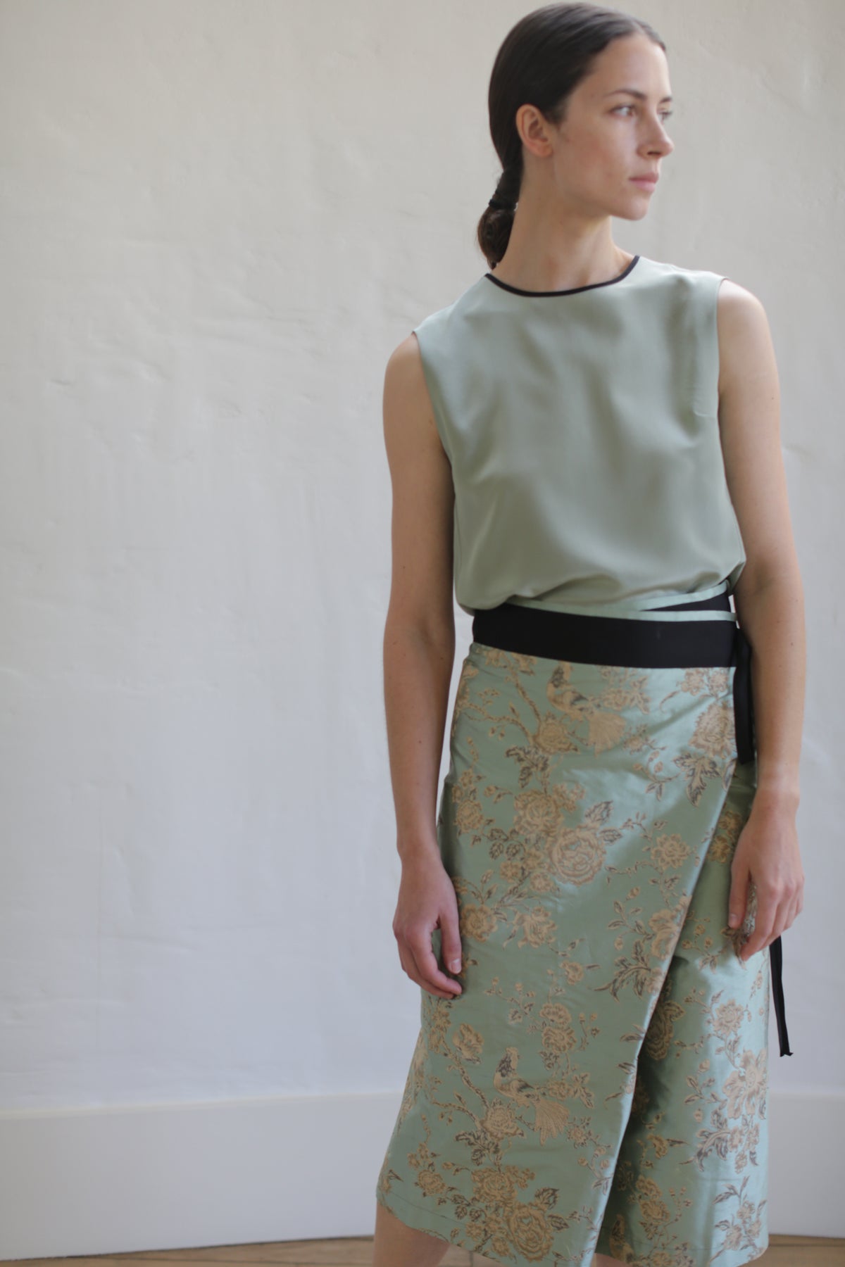 A woman in a sleeveless green top and the limited edition Dupioni Petal Wrap Skirt in Jade stands against a light-colored wall. Her hair is tied back, and she gazes thoughtfully to the side.