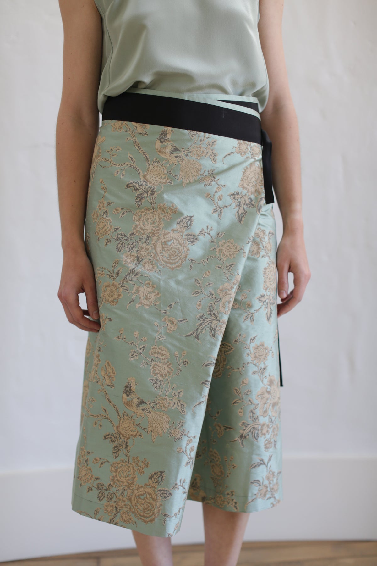 The person is wearing the Dupioni Petal Wrap Skirt in jade, featuring a floral pattern and a black waistband, crafted from textured Dupioni silk. They pair it with a matching sleeveless top. The photo showcases this limited edition ensemble indoors against a white background.