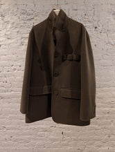 Load image into Gallery viewer, The Sculptor&#39;s Jacket in olive, featuring three buttons, flap pockets, and a notched collar, hangs on a white hanger against a textured white brick wall.