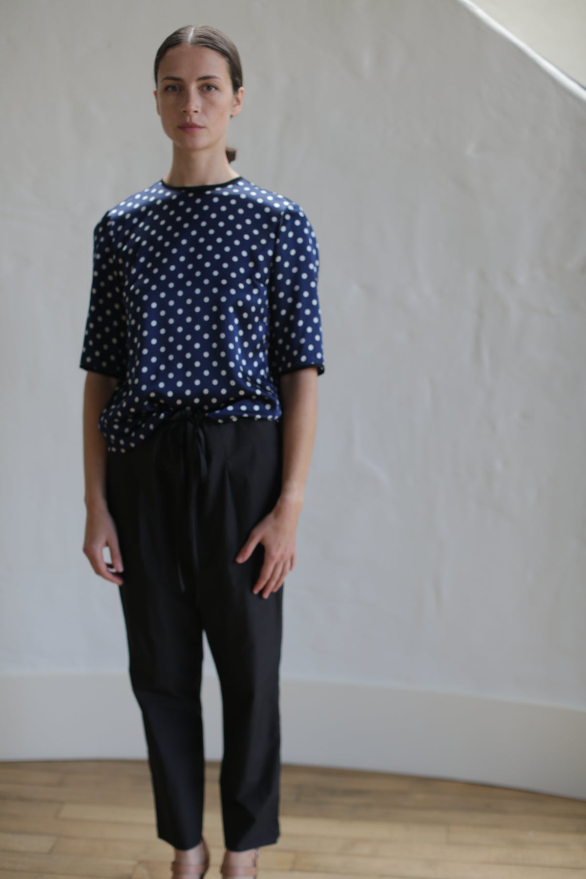 A person stands indoors against a plain, light-colored wall. They are wearing a navy blue shirt with white polka dots and Typewriter Unisex Pants | Black. The person has a neutral expression and their hair is pulled back with a Japanese grosgrain ribbon. The floor appears to be wooden.