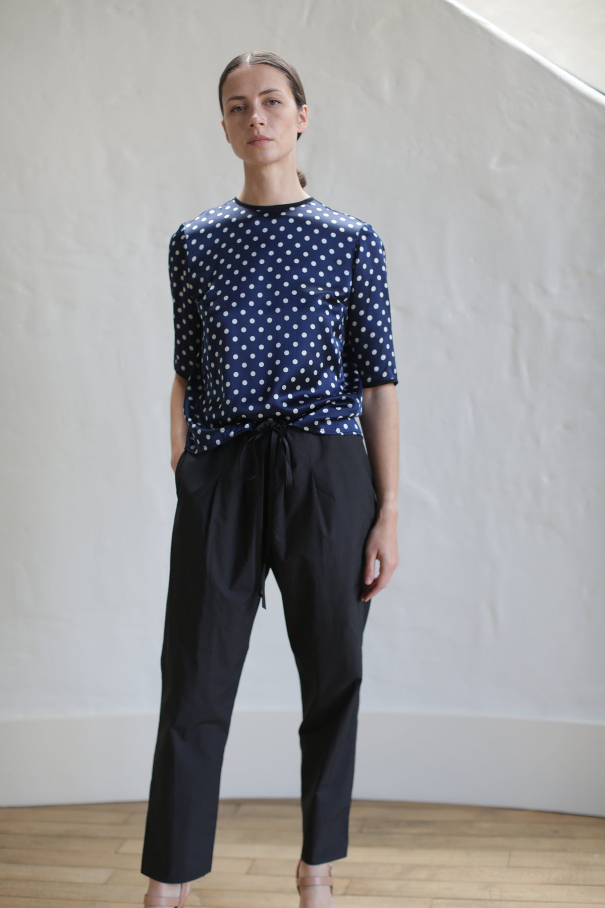 A person is standing indoors against a light gray wall, dressed in a navy blue shirt with white polka dots and Typewriter Unisex Pants | Black. They have their hands in their pockets and are looking directly at the camera with a neutral expression.