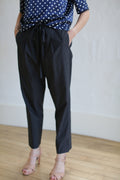 A person is standing against a plain white wall on a wooden floor, wearing a navy polka dot blouse accented with Japanese grosgrain ribbon, Typewriter Unisex Pants | Black, and nude-colored open-toe heels. Their hands are in their pockets, and their face is not visible in the image.