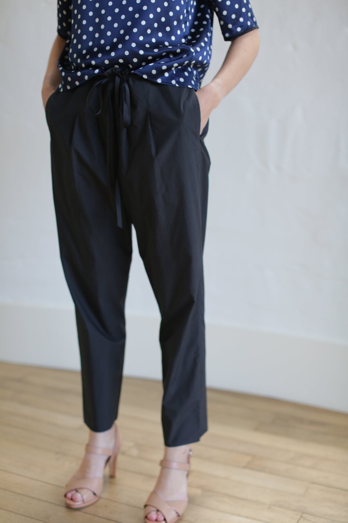 A person is standing against a plain white wall on a wooden floor, wearing a navy polka dot blouse accented with Japanese grosgrain ribbon, Typewriter Unisex Pants | Black, and nude-colored open-toe heels. Their hands are in their pockets, and their face is not visible in the image.