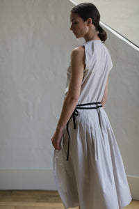 A woman stands in a softly lit room with a neutral-colored wall. She is wearing the Typewriter Wrap Dress | Dove, crafted from Typewriter Cotton with pleats and a black ribbon belt around her waist. Handmade in San Francisco, her hair is tied back as she gracefully faces away, slightly looking to the side.