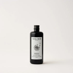 A black bottle with a metallic screw cap boasts a minimalist label design. "Wilder Botanicals" is written at the top of the label, with "Immortelle Cleansing Oil" at the bottom. The central part of the label showcases a floral illustration, accentuating its blend of skin-renewing botanicals such as organic Manuka. The background is plain white.