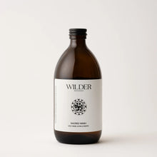 Load image into Gallery viewer, A brown glass bottle with a black cap labeled &quot;Sacred Wash&quot; by Wilder Botanics sits on a white background. The label features a circular floral design and text that mentions &quot;Holy Basil &amp; Palo Santo extract.&quot; This hand and body wash boasts a minimalist, natural aesthetic.