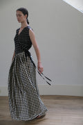 A woman with dark hair is standing in a light-filled room, wearing the Charmeuse Wrap Dress | B/W Polkadot. The dress has a fitted top and a flowing wrap skirt, accentuated with a black ribbon tied at the waist. She is looking downward with a serene expression.