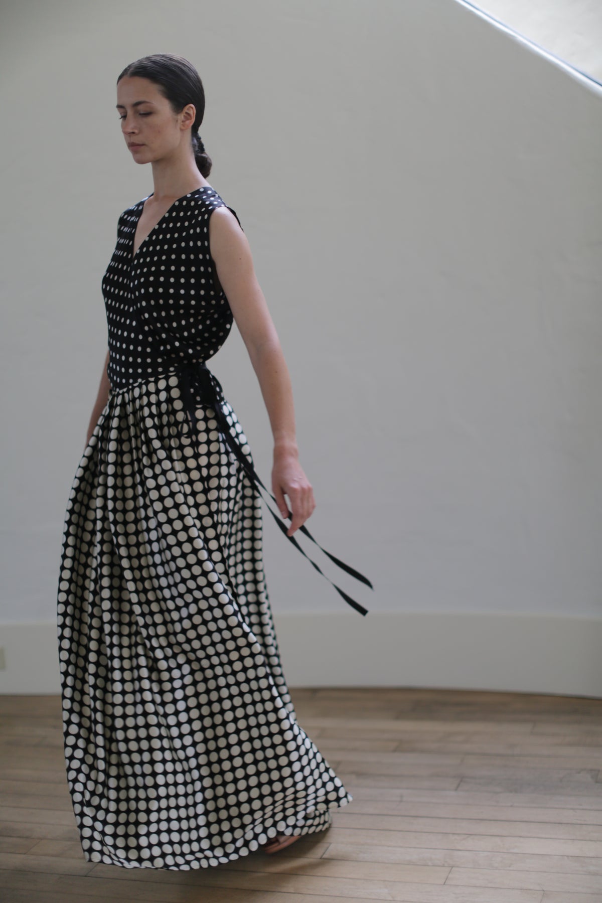 A woman with dark hair is standing in a light-filled room, wearing the Charmeuse Wrap Dress | B/W Polkadot. The dress has a fitted top and a flowing wrap skirt, accentuated with a black ribbon tied at the waist. She is looking downward with a serene expression.
