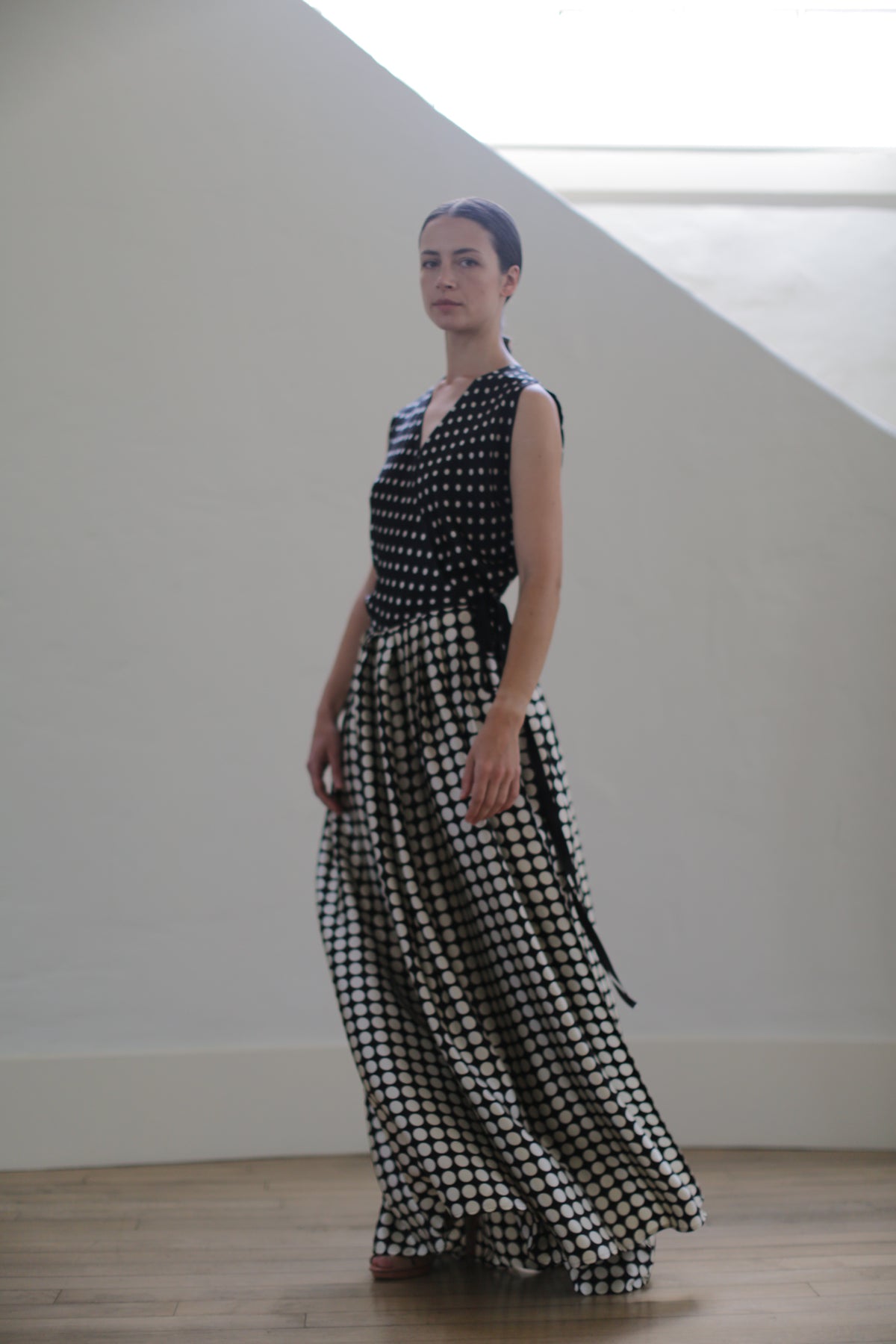 A person stands indoors wearing a Charmeuse Wrap Dress | B/W Polkadot. Made from charmeuse silk, the upper part of the dress is fitted, switching to a flowing wrap skirt. They have dark hair pulled back and are positioned against a light-colored wall.