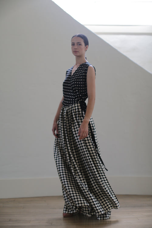 A person stands indoors wearing a Charmeuse Wrap Dress | B/W Polkadot. Made from charmeuse silk, the upper part of the dress is fitted, switching to a flowing wrap skirt. They have dark hair pulled back and are positioned against a light-colored wall.
