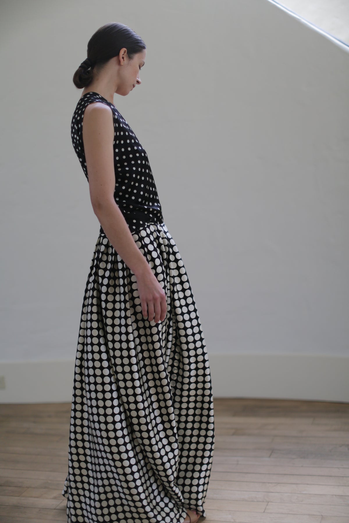 A person stands in a minimalist, well-lit room wearing a Charmeuse Wrap Dress | B/W Polkadot. They face slightly downward with their hair tied back in a low bun. The room features wooden flooring and a plain backdrop.