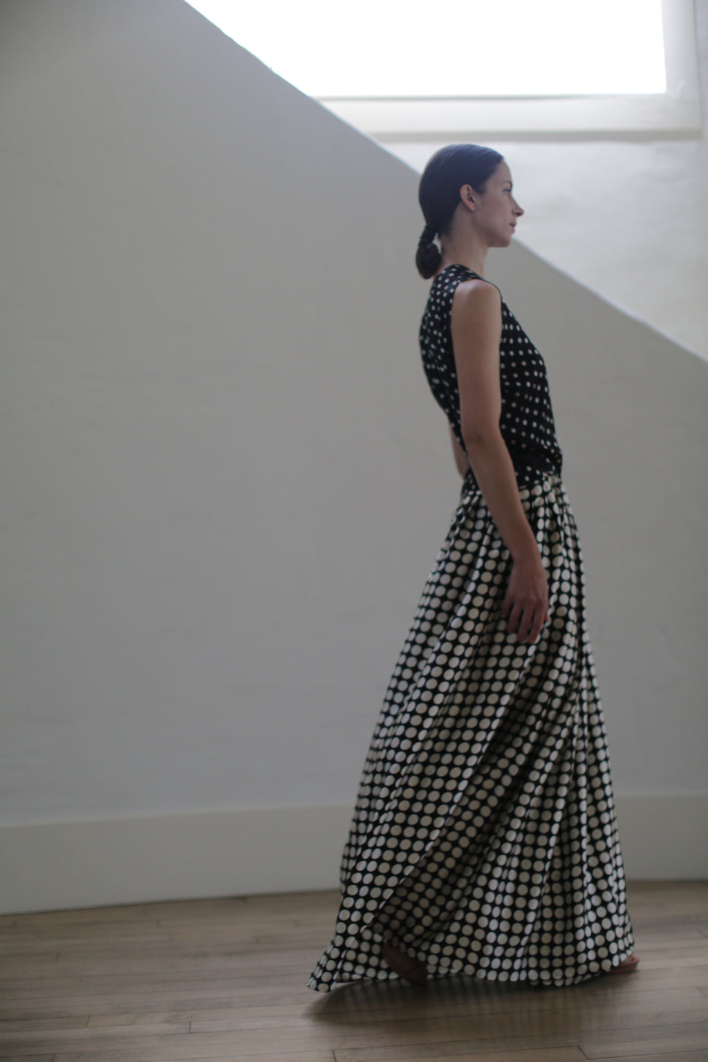 A woman in profile walks indoors wearing a Charmeuse Wrap Dress | B/W Polkadot. Her hair is tied back, and she is against a plain white wall with light coming from a window above. The room has light wood flooring.