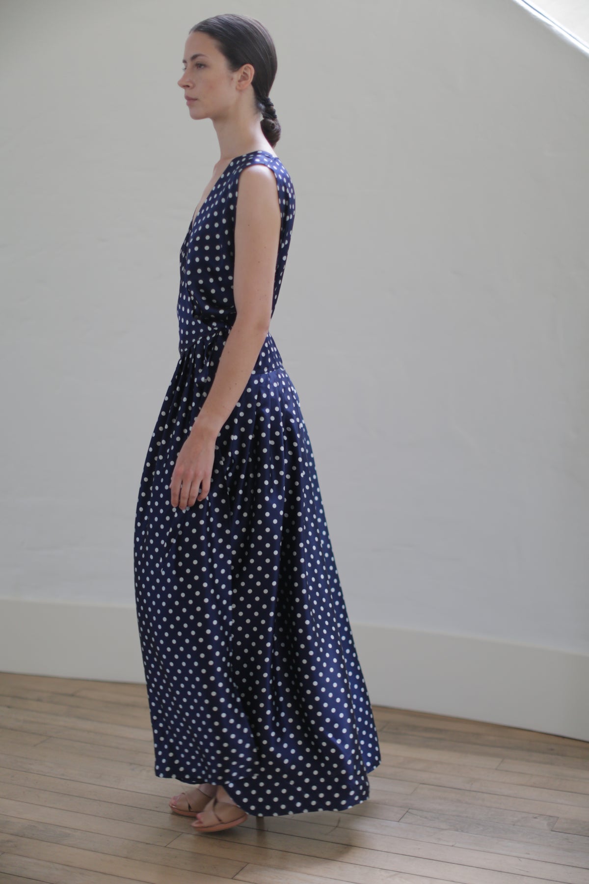 A woman stands in profile, wearing a sleeveless Charmeuse Wrap Dress | Navy Polkadot. Her dark hair is pulled back into a low ponytail, secured with Japanese silk grosgrain straps. She stands on a wooden floor in front of a plain, light-colored wall.